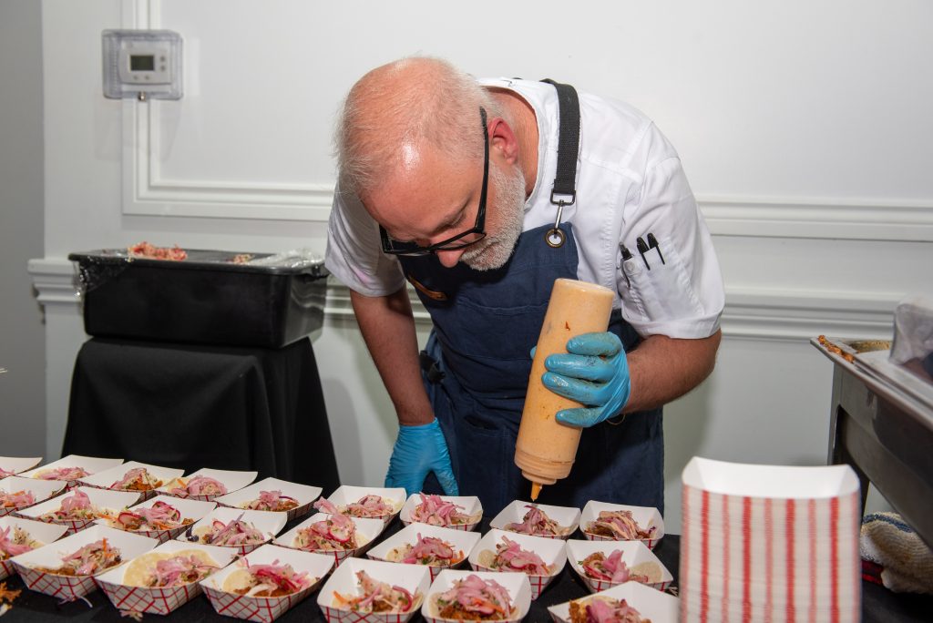 A chef wearing glasses and blue gloves, leaning over a table to drizzle sauce onto small, individually portioned dishes.