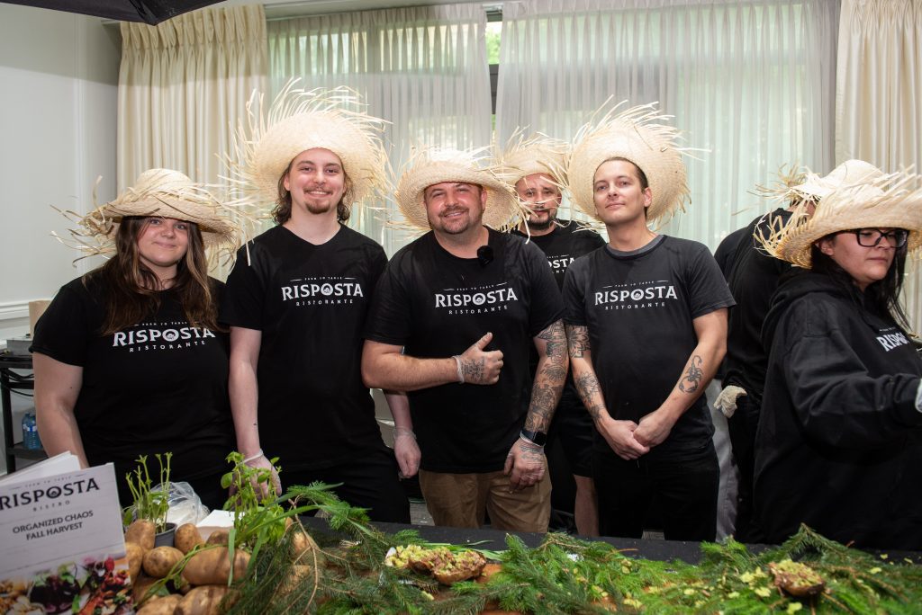 A group of six chefs wearing black t-shirts that say "RISPOSTA RISTORANTE" and straw hats, standing behind a food display.
