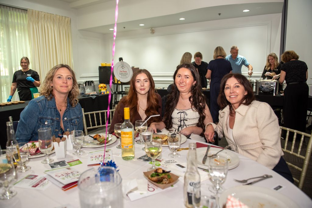 Four women seated at a dining table with food and drinks, smiling, in a brightly lit event room.